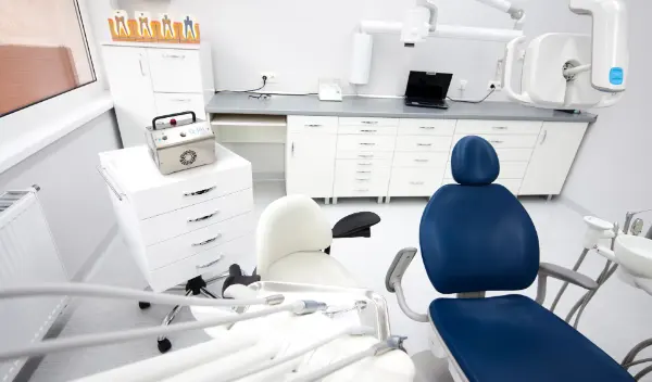 Zahnarztpraxis mit blauem Behandlungsstuhl, weißem Stuhl, weißen Schränken und zahnärztlichen Geräten.