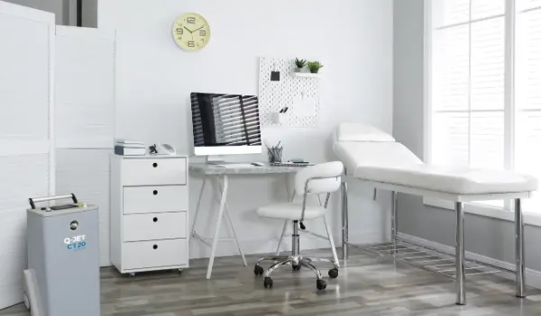 Helle Arztpraxis mit Untersuchungsliege, Schreibtisch, Computer, Stuhl auf Rollen, Schubladenschrank und Uhr an der Wand.