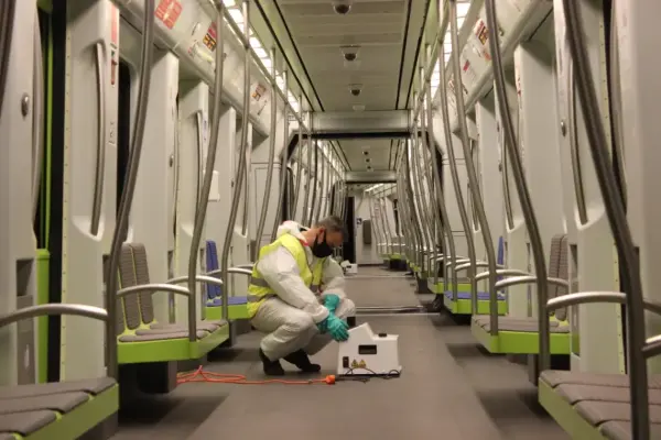 Person in Schutzanzug und Warnweste kniet in einem leeren, modernen U-Bahn-Wagen mit grünen Sitzflächen und grauem Boden und bedient ein weißes Gerät.