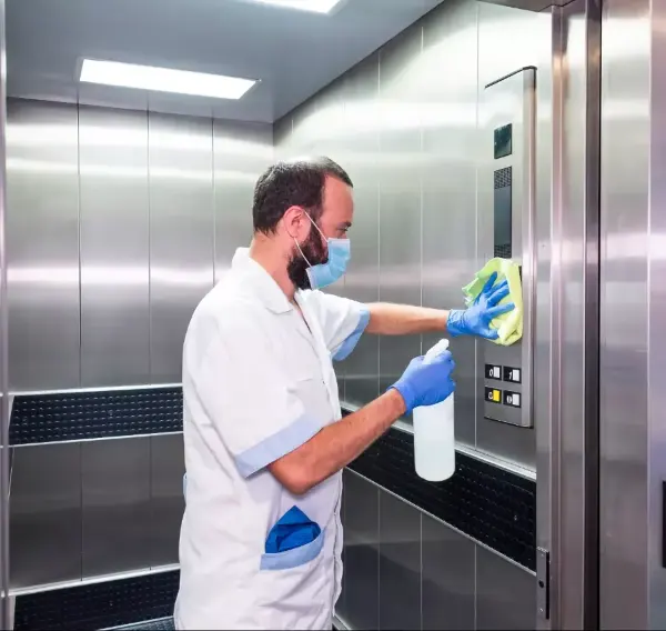 Mann in weißer Arbeitskleidung reinigt mit Sprühflasche und Tuch die Innenseite einer Aufzugtür aus Edelstahl.