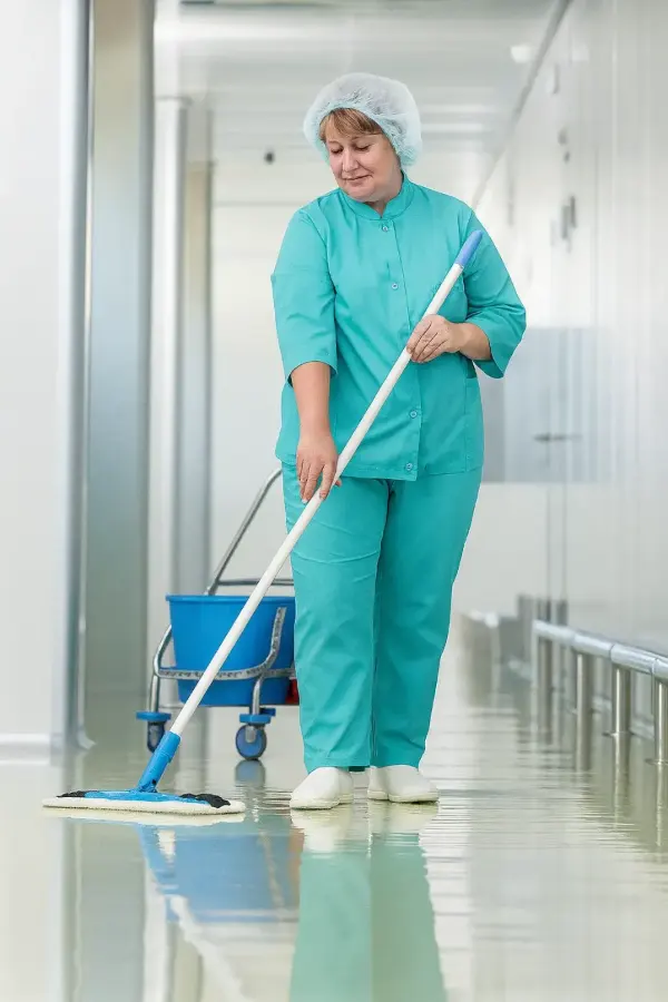 Person in medizinischer Schutzkleidung und Haube wischt mit Wischmopp einen glänzenden Flur, blauer Eimerwagen im Hintergrund.