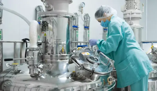 Person in Schutzkleidung bedient großes Edelstahl-Laborgerät mit Ventilen und Rohren in einem Labor.