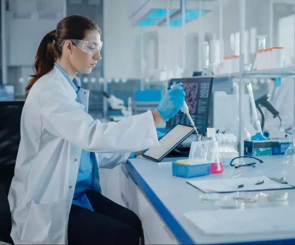 Laborantin in weißem Kittel und blauer Handschuh hält Pipette über Reagenzgläser auf Labortisch mit Monitor und Laborutensilien.