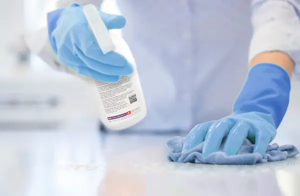Person mit blauen Handschuhen sprüht Desinfektionsmittel aus einer weißen Sprühflasche auf eine Oberfläche und wischt sie mit einem blauen Tuch ab.