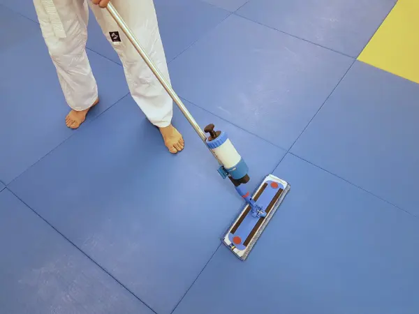 Barfüßige Person in weißem Anzug wischt blauen Boden mit langem Wischmopp, gelber Bereich am Rand sichtbar.