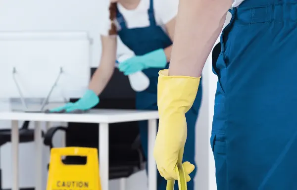 Zwei Reinigungskräfte in blauen Overalls und gelben Handschuhen reinigen ein Büro mit Schreibtischen und Computern, Warnschild steht auf dem Boden.