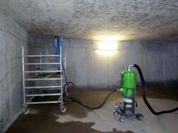 Person in blauer Schutzkleidung steht auf einem fahrbaren Gerüst in einem kahlen Betonraum mit einer grünen Industriestaubsaugeranlage.