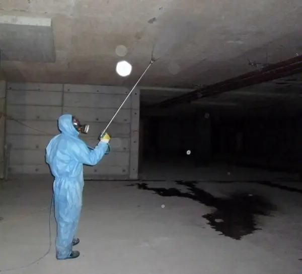 Person in blauem Schutzanzug und Atemmaske hält Stab in leerem, dunklem Betonraum mit dunklem Fleck auf dem Boden.