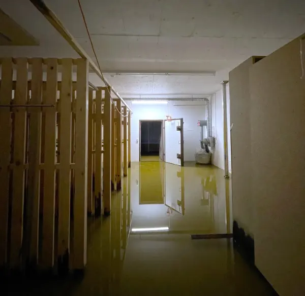 Flur mit Holzregalen und offener Tür, Boden steht unter stehendem Wasser, Decke mit sichtbaren Rohren und Kabeln.