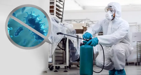 Person in weißem Schutzanzug, Atemschutzmaske und Schutzbrille sprüht Desinfektionsmittel in einem Raum mit Regalen. Ein vergrößerter blauer Virus mit durchgestrichenem Kreis symbolisiert Desinfektion.